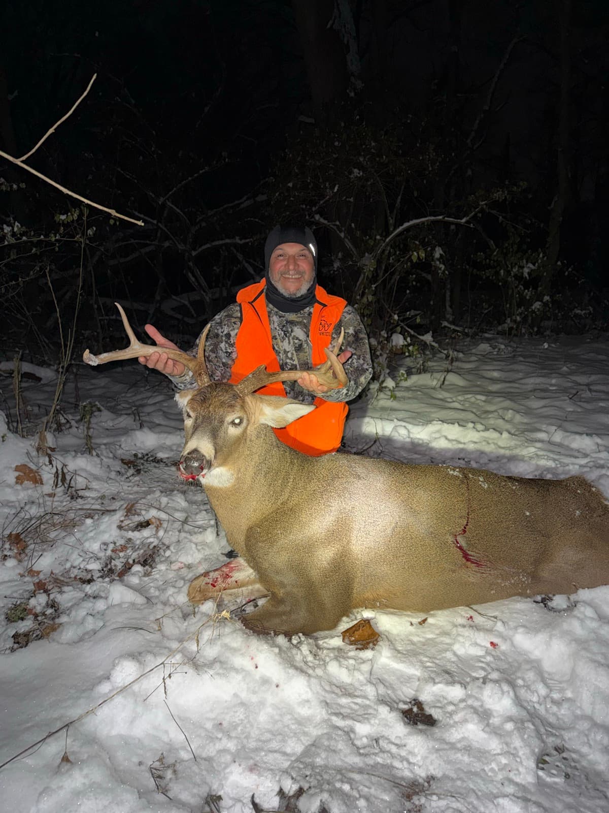 Hunter in a blaze orange vest with a buck recovered in the snow at night